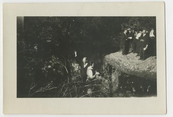 Unidentified man performing a baptism on an unidentified woman in a stream while a crowd watches from shore