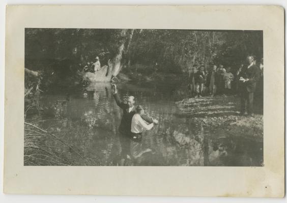 Unidentified man baptizes an unidentified man in a stream while a crowd watches from shore