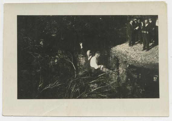 Unidentified man baptizes an unidentified man in a stream while a crowd watches from shore