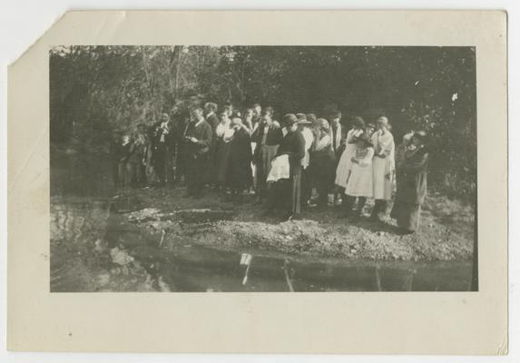 Unidentified crowd standing on shore of a stream
