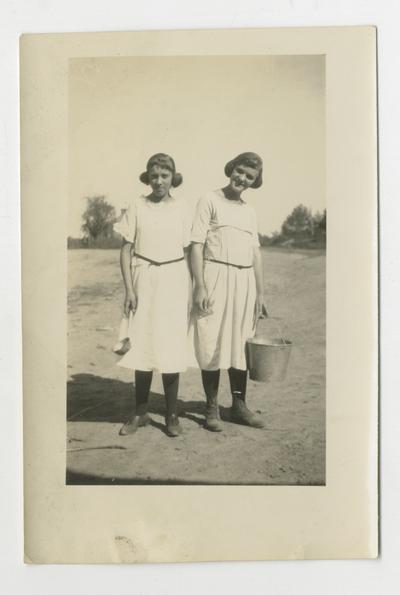 Two unidentified young women, one holding a ladle and other holding a bucket