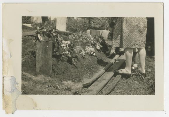 Unidentified persons (from leg-down) standing in front of flowers on top of freshly covered gravesite