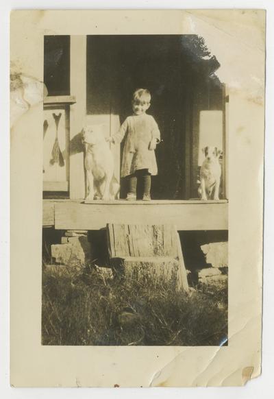 Unidentified young child on porch with a dog on either side of the child