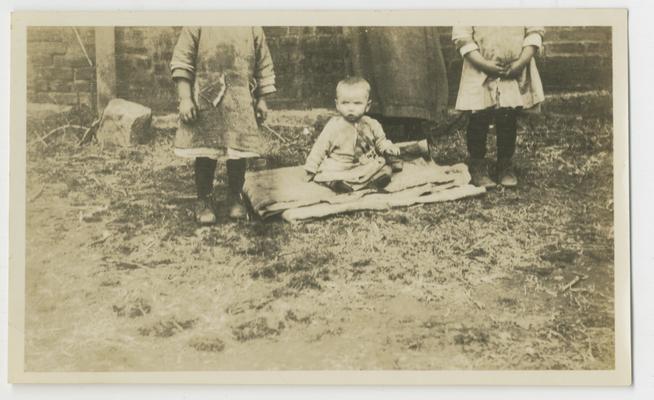 Unidentified infant sitting on blanket on the ground with three unidentified persons in background, cutoff by image