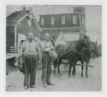 Two workmen standing by two horses hitched to a wagon