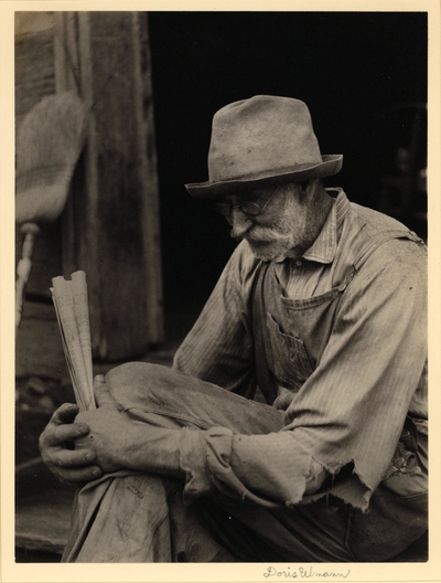 Elderly, bearded man in hat, glasses, ragged shirt, and overalls, seated, holding rolled-up newspaper; broom leaning against wall