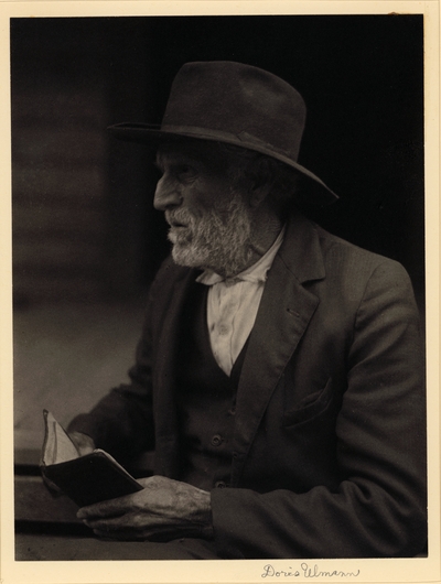 Elderly, bearded man in hat and suit, seated with small book