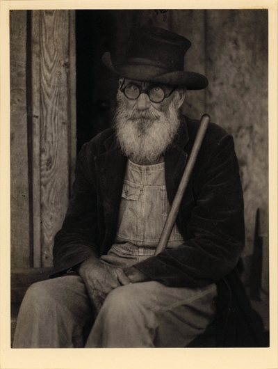 Elderly, bearded man in hat, glasses, coat, and overalls, seated with broomstick