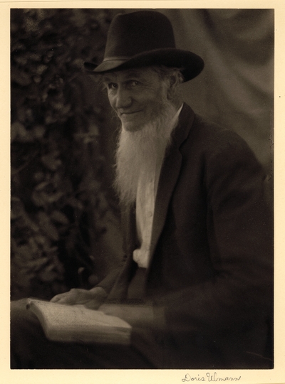 Elderly, bearded man in hat and coat, seated with book