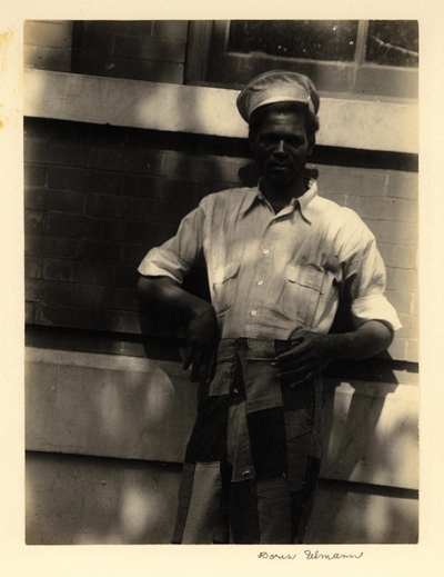 Black man in hat, shirt with rolled-up sleeves, and patched pants, leaning against wall of brick building