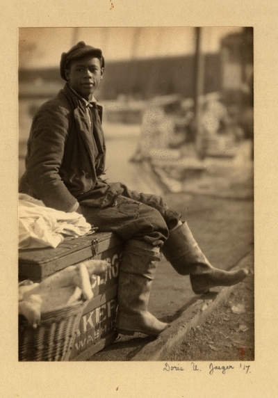 Young black man in hat, coat, patched pants, and boots, seated on trunk with basket beside it.  1917