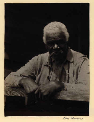 Elderly black man with mustache, in ragged coat, seated behind wooden rail, with hands on rail