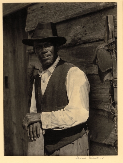Bearded black man in hat and vest, standing with hands on post, in front of wooden building with draft horse bridle hanging on peg