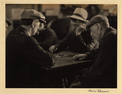Three men in hats and coats, seated around table, playing checkers, with other men in background