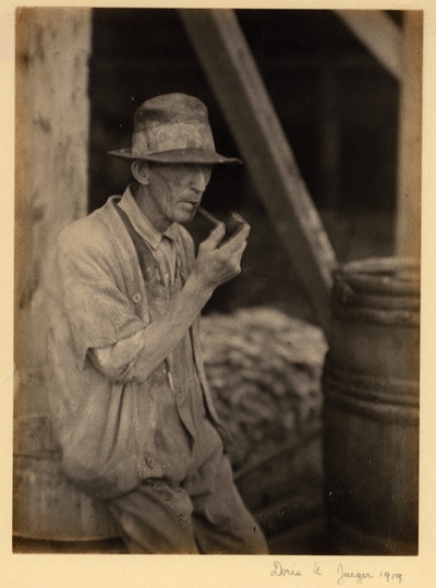 Elderly man in ragged hat, sweater, and overalls, leaning against barrel, smoking pipe with barrel beside him and beams in background