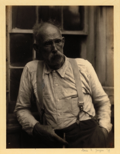 Elderly man with mustache and suspenders, leaning against wall with window, holding pipe, with other hand in pocket