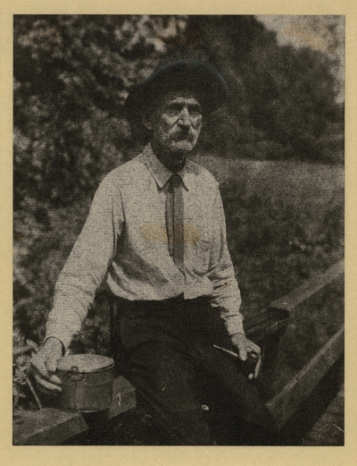 Elderly man with mustache, in hat, shirt, and tie, seated on fence, holding pipe and small pail, with trees in background