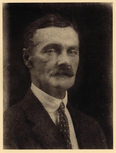 Head shot of man with mustache, in coat and polka-dot tie