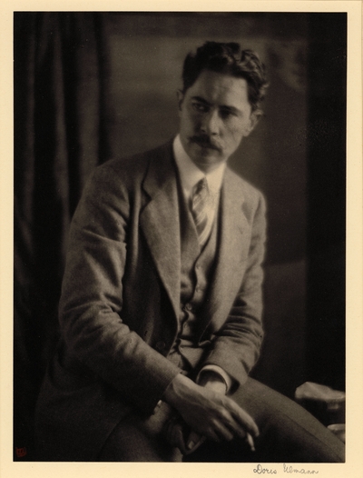 Man with mustache, in suit and tie, seated, holding cigarette