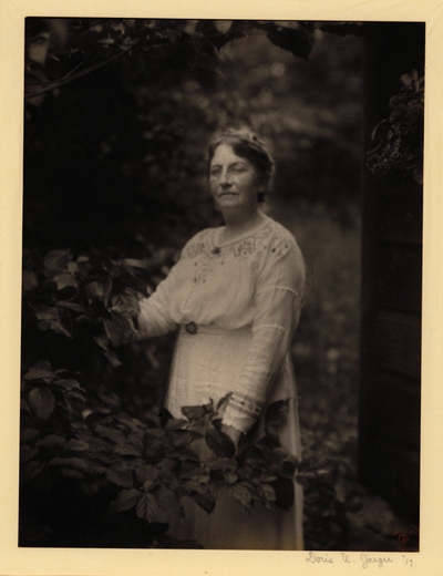 Woman in glasses and white dress, standing behind branches of tree, with her hands on them