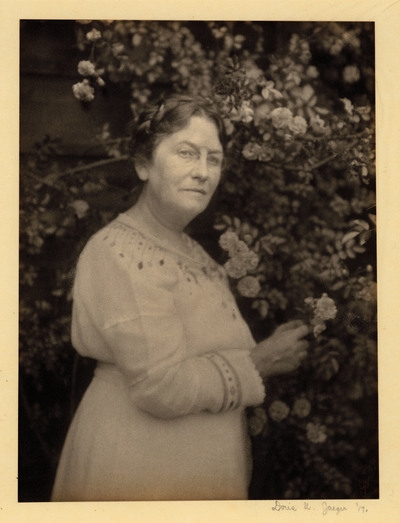 Woman in glasses and white dress, standing beside flowering shrub, touching a branch