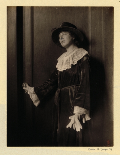 Woman in hat and velvet dress with lace collar and cuffs, standing in front of door, holding gloves