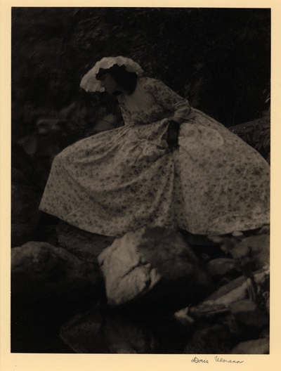 Profile of woman in lace hat, full dress, and gloves, sitting on rocks