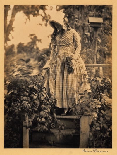 Young woman in bonnet and dress, holding flowers, standing on steps surrounded by plants, with birdhouse in background