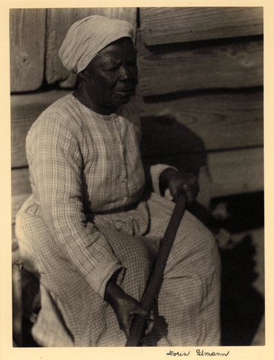 Black woman in turban and dress, seated against wood building, holding long wooden handle