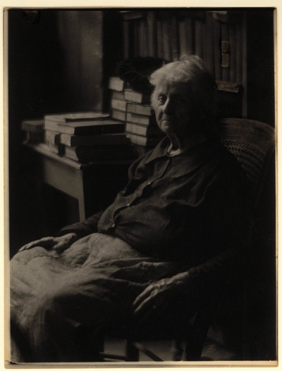 Elderly woman in blouse and skirt, seated in chair, with bookshelf and table with books and hat in the background