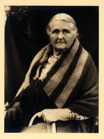 Elderly Native American woman with blanket draped over her shoulders, seated with basket handle showing