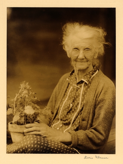 Elderly woman in glasses, dress, and sweater, seated, holding potted plant in her lap
