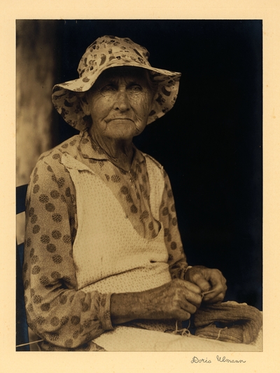 Elderly woman in hat and dress with apron, seated in chair with sweater and yarn in her lap, knitting