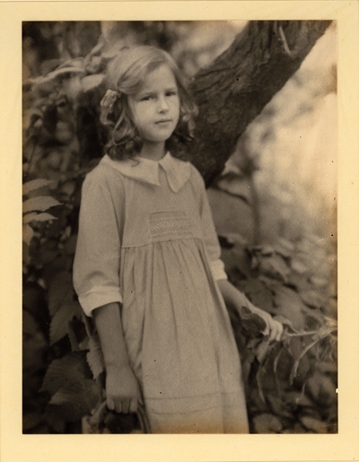 Girl in ringlets, ribbon, and dress, standing in front of tree