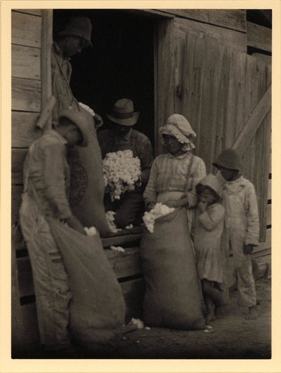 Barefoot boy and girl standing behind woman and three men in doorway with bags of cotton