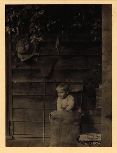 Small child seated in chair on porch with broom, hat, and hanging plants against wall