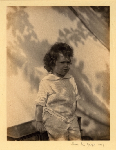 Small, curly-haired child standing in front of drape with shadows of leaves across it