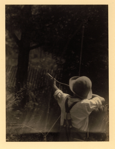 Back view of boy in hat and overalls, aiming homemade bow and arrow, with tree in background