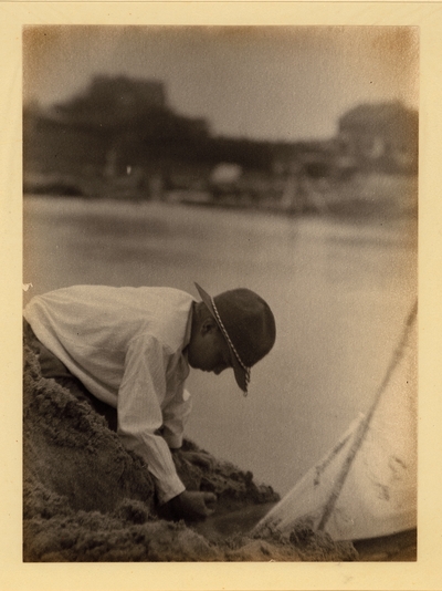 Profile of boy in hat digging in sand on the edge of water, with sailboat beside him.  1918-1921