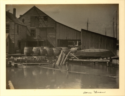 Weathered buildings on wharf, with barrels in front and masts of ships showing in background