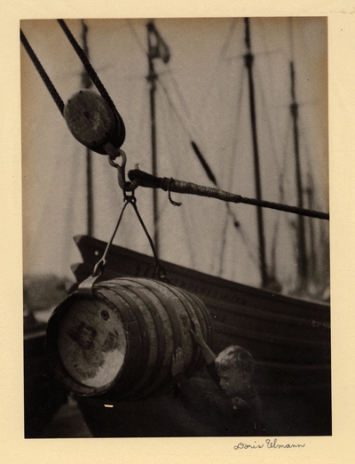 Barrel suspended on hook with boy steadying it and ships in background