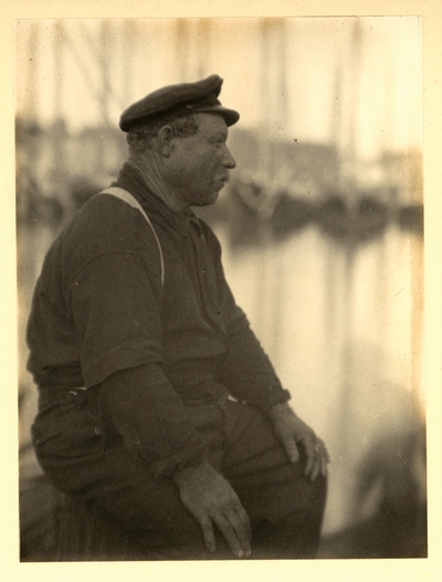 Profile of man in hat and suspenders, seated with ships in background