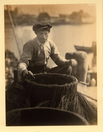 Man with mustache, in hat and striped shirt, behind barrel with lines running into it