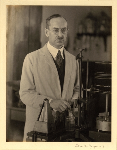 Man in glasses, lab coat, vest, and tie, standing in lab with metronome and other apparatus, holding reagent bottle