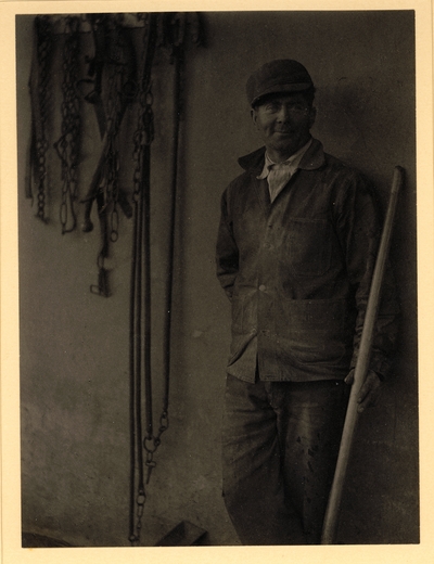 Man in hat and coat, holding long handle, leaning against wall with harness hanging from it