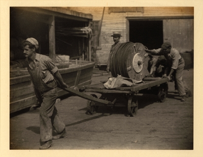 One man pulling and several others pushing wagon with large spool, with building and doorway in background