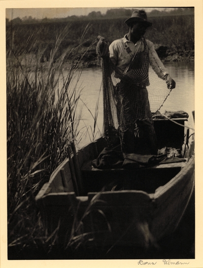 Black man in hat and overalls, holding net, standing in boat on small waterway surrounded by tall grass.  South Carolina, ca. 1929-31