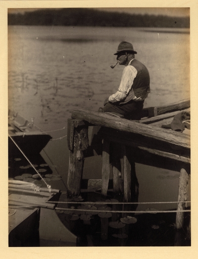 Elderly man with hat and pipe, seated on dock, fishing