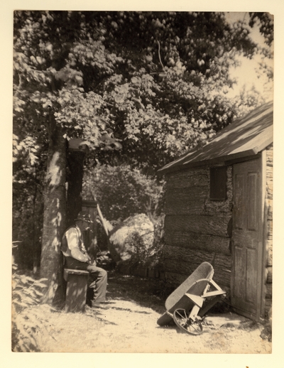 Elderly man with hat and pipe, sitting on bench beside log cabin and overturned wheelbarrow