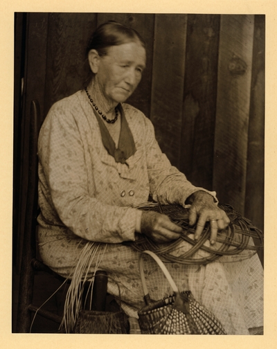 Aunt C. Ritchie;  Basket-maker, Hindman, KY; Elderly woman in dress and necklace, seated in chair, making basket with baskets beside her